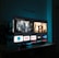 A television screen displaying a user interface with popular streaming service apps including Netflix, Sky, Apple TV, Prime Video, and others. The screen also shows images from movies or shows above the apps. The room is dimly lit with a potted plant visible on the side.