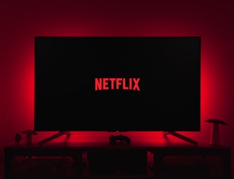A dark room featuring a television screen displaying the word 'Netflix' in red against a black background. The screen is centered on a console table, with a video game controller placed in front. The room is dimly lit by red ambient lighting, creating a cozy atmosphere.