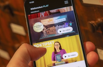 A person holding a smartphone displaying a webpage with a Scandinavian library streaming service. The screen shows two video thumbnails, one with a TV displaying a program and the other with a woman holding a book. The background features a wooden drawer set with labels.