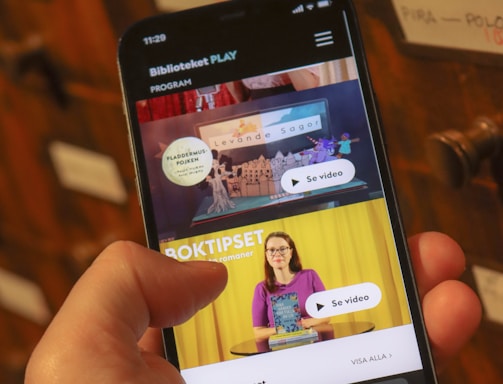 A person holding a smartphone displaying a webpage with a Scandinavian library streaming service. The screen shows two video thumbnails, one with a TV displaying a program and the other with a woman holding a book. The background features a wooden drawer set with labels.
